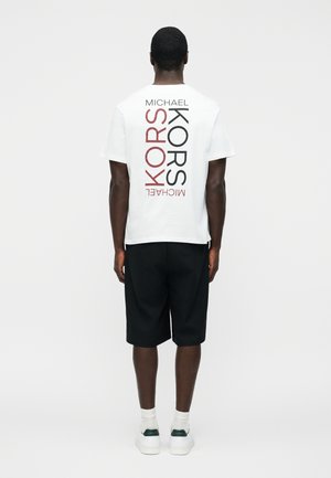 Άνδρας που φοράει λευκό T-shirt Michael Kors με κατακόρυφο κόκκινο και μαύρο λογότυπο στο πίσω μέρος, μαύρα σορτς, λευκές κάλτσες και λευκά αθλητικά παπούτσια.