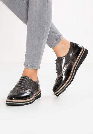 Sorte og metaliske wingtip oxford-sko med lagdelt sål båret med lysegrå skinny jeans mod en hvid baggrund.