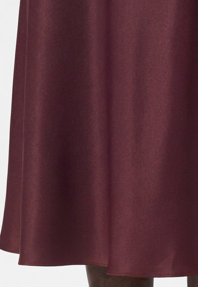 Κοντινό πλάνο στο κάτω μισό μιας μπουρντό σατέν φούστας που φοράει ένα άτομο με σκούρο δέρμα, παρουσιάζοντας την υφή του υφάσματος και τη λεπτομέρεια του ποδόγυρου.