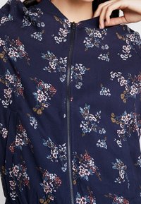 Granatowa kurtka z zamkiem z przodu, elastycznym pasem i małym, wielokolorowym kwiatowym wzorem, noszona przez osobę z ręką blisko kołnierza.