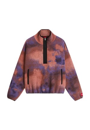 Person trägt lila und pinken Fleece-Pullover, lockere schwarze Hose, schwarze Sneaker, Sonnenbrille, hält eine bronzefarbene Steppjacke und eine gelbe Umhängetasche.
