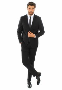 Abito da uomo nero con vestibilità su misura, camicia bianca e cravatta nera. Scarpe eleganti nere lucide. Postura in piedi con una mano in tasca.