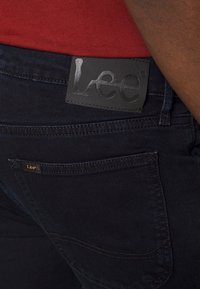 Mörkblå denimjeans med en svart läderlogga på midjan, accentuerad med guldtråd och en liten logglabel på bakfickan.