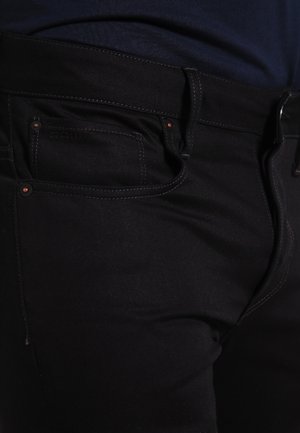 Zbliżenie kieszeni czarnych dżinsów z widocznym przeszyciem i miedzianymi nitami na osobie ubranej w ciemną koszulę.