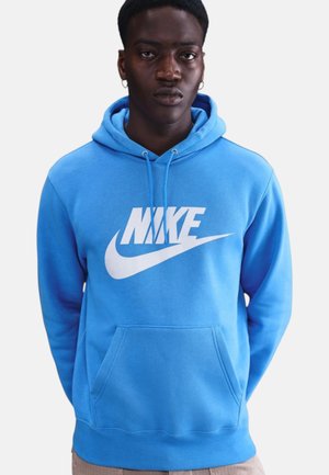 Uomo che indossa una felpa Nike blu brillante con logo bianco, in piedi contro uno sfondo chiaro uniforme, con le mani dietro la schiena.