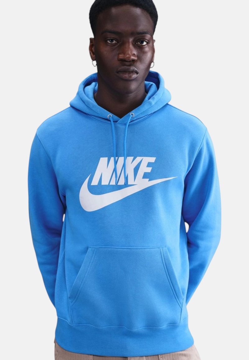 Uomo che indossa una felpa Nike blu brillante con logo bianco, in piedi contro uno sfondo chiaro uniforme, con le mani dietro la schiena.