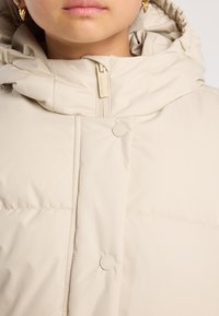 Primo piano di una persona che indossa una giacca imbottita beige con colletto alto e cappuccio, zip e bottoni a pressione, che mostra la parte bassa del volto e orecchini d'oro.