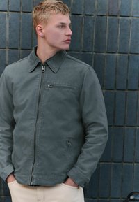 Veste grise zippée avec un col en velours côtelé, des poches latérales et de poitrine, et une texture lisse, portée sur un pantalon de couleur claire.