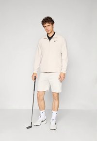 Svetlo bež jopica z zadrgo, črna srajca z ovratnikom, bele kratke hlače in beli supergi. Drži golf palico, z enobarvno ozadje.