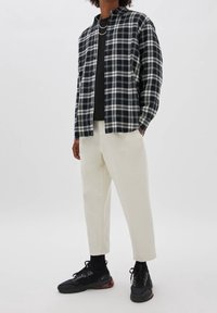 Persona che indossa una camicia a quadri bianco e nero sopra una maglietta nera, pantaloni corti color avorio, scarpe da ginnastica nere con accenti rossi e collane a catena d'argento.