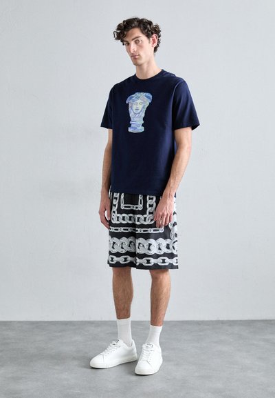 T-shirt bleu marine avec un graphique d'une tête sculptée ; shorts noirs avec un motif de chaîne métallique ; baskets blanches ; matériau en coton lisse.