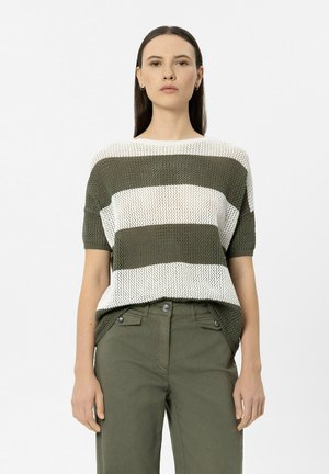 Frau trägt ein grün-weiß gestreiftes Strick-Shortsleeve-Oberteil und olivgrüne, hoch taillierte Hose vor einfarbigem Hintergrund.