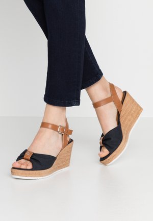 Piedi con sandali zeppa in tessuto nero e pelle color cuoio con cinturini alla caviglia, indossati con pantaloni corti blu scuro su pavimento chiaro.
