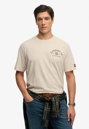 Hombre con camiseta beige de Superdry, jeans azules, camisa de cuadros atada a la cintura y reloj, de pie con una mano en el bolsillo sobre fondo blanco.