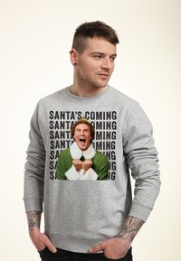 Szary sweatshirt z grafiką na przodzie, na której znajduje się napis "ŚWIĘTY MIKOŁAJ NADCHODZI" oraz obrazek postaci ubranego w zielony strój z białymi akcentami.