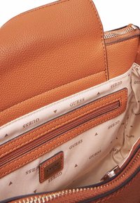 Interno di una borsa a tracolla in pelle color cuoio con una tasca con zip, caratterizzato da una fodera beige stampata con "GUESS" e piccoli motivi a triangolo.
