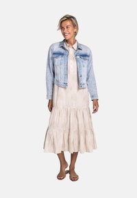 Jeansjacke, hellblau, in verkürztem Design. Darunter ein beiges Kleid mit gestuftem Saum und dezentem abstraktem Muster. Kombiniert mit neutralen Sandalen.