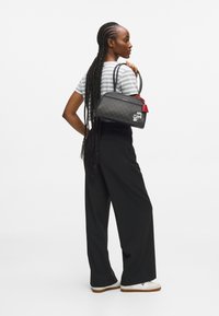 Geantă neagră cu o suprafață cu model, accent roșu și etichetă decorativă. Model îmbrăcat cu o bluză cu dungi și pantaloni negri larghi, întors cu spatele.