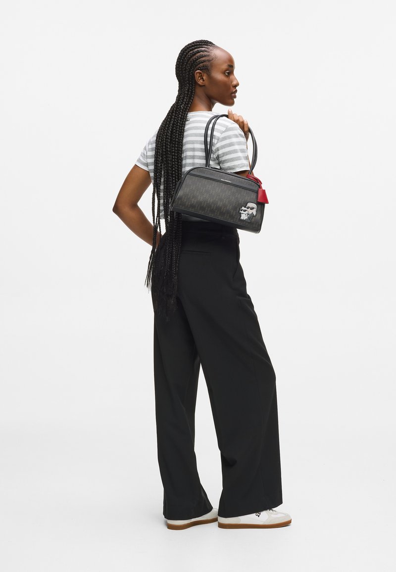 Czarna torebka z wzorzystą powierzchnią, czerwoną akcentacją i dekoracyjną etykietą. Modelka nosi paskowany t-shirt oraz szerokie czarne spodnie, zwrócona plecami.