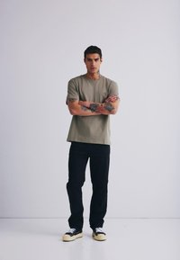 Olivno zelena majica s kratkimi rokavi in žepom na prsih, v kombinaciji s črnimi kavbojkami in črnimi supergami z belimi vezalkami. Model stoji s crossed arms.