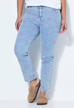 Personne portant un jean bleu clair avec des motifs de soleil roses et des chaussures marron sans lacets, debout contre un mur blanc.