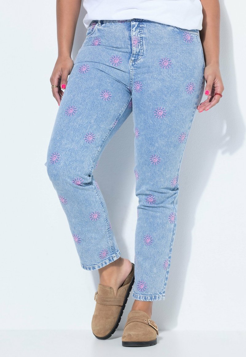 Personne portant un jean bleu clair avec des motifs de soleil roses et des chaussures marron sans lacets, debout contre un mur blanc.