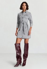 Abito camicia in denim grigio con chiusura a bottoni sul davanti, maniche lunghe e cintura regolabile. Indossato con stivali al ginocchio lucidi color bordeaux scuro.