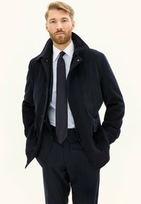 Tummansininen takki, jossa on kohotettu kaulus, pehmeää kangasta. Varustettu vetoketjulla, sivutaskuilla ja muotoilultaan istuva, yhdistettynä raidalliseen paitaan ja solmusidokseen.