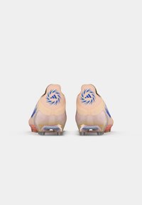 Un par de botas de fútbol color durazno con logotipos en espiral azules en los talones y suelas translúcidas con tacos naranjas vistas desde la parte trasera.