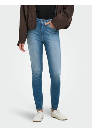 Personne portant un jean skinny bleu, des baskets blanches, un haut noir et une veste marron, avec une main dans la poche, sur un fond clair uni.