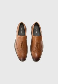 Zapatos de cuero marrón tipo slip-on con un diseño texturizado, borlas decorativas, detalles de brogue y una punta redondeada elegante.