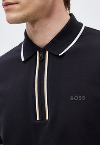 Homme portant un polo noir avec un bord blanc sur le col et une fermeture éclair à rayures verticales beige et blanche, logo "BOSS" sur la poitrine.