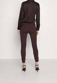 Brązowe, dopasowane spodnie o gładkiej fakturze z zamkiem błyskawicznym po bokach, w połączeniu z ciemną, długą koszulą z guzikami i sandałami na obcasie.