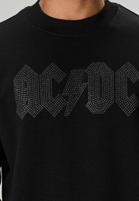Fekete pulóver bordázott nyakkivágással, melyen feltűnő, rhinestone stílussal díszített felirat olvasható: "AC/DC", valamint egy cikcakkos villám mintázat.