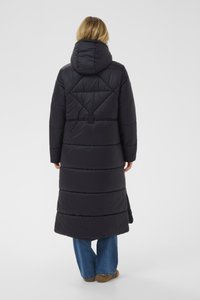 Melna polsterēta gara jaka ar kapuci, ar ģeometriska raksta šūšanu, sānu šķēlumu un teksturētu apdari. Kombinēta ar zilajiem džinsiem un brūnām kurpēm.