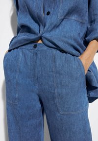 Sininen chambray-paitapusero nappilistan ja rennon leikkauksen kanssa, yhdistettynä samaan sävyyn löysiinkäyttäviin housuihin, joissa on läppätaskut ja ommellut yksityiskohdat.