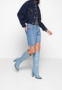 Vaaleansinisiä, polven korkeudelle ulottuvia saappaita, joissa on kiiltävä, teksturoitu pinta ja paksu korko. Yhdistetty farkkutakin ja repaleisten farkkushortsejen kanssa.