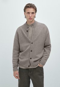 Jasno brązowy sweter z dzianiny z wysokim kołnierzem, zapinany na guziki, z prążkowanym pasem, noszony na beżowym swetrze i ciemnych dżinsach.