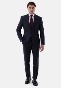 Marineblauer Anzug mit tailliertem Schnitt, ausgestattet mit einem einreihigen Knopfverschluss, Revers mit Einkerbung und Seitentaschen, kombiniert mit einem weißen Hemd und einer dunkelroten Krawatte.