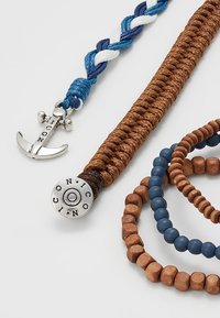 Köynnöksinen sinivalkoinen köysi-rannekoru, jossa on hopea ankkurihelmi, yhdessä ruskeiden kudottujen ja helmillä varustettujen rannekorujen kanssa, jotka vaihtelevat koossa ja tekstuurissa.