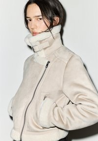 Beżowa kurtka z materiału imitującego owczą skórę, z wysokim kołnierzem, zamkiem błyskawicznym z przodu oraz miękkimi białymi futrzanymi akcentami przy mankietach i kołnierzu. Posiada boczne kieszenie oraz teksturowane wykończenie.