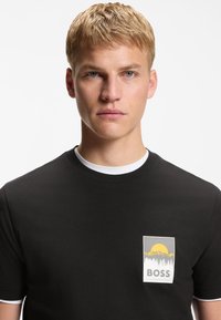 Νεαρός άνδρας με ξανθά κοντά μαλλιά που φοράει μαύρο T-shirt με κίτρινο σχέδιο ήλιου, βουνού και δέντρου και τη λέξη «BOSS» στο στήθος.