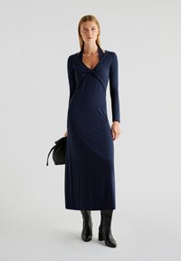 Navy blauwe jurk met lange mouwen en een crossover voorkant, verlengende silhoët, zachte stof en een subtiele glans, gecombineerd met zwarte enkellaarsjes en een tas.