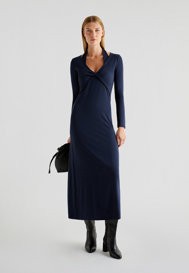 Robe marine à manches longues avec un devant croisé, silhouette élancée, tissu doux et un léger éclat, assortie à des bottines noires et un sac.