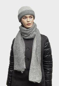 Donna con un berretto di lana grigio e una sciarpa larga a righe grigie sopra un cappotto nero con maniche imbottite, che guarda leggermente a destra su uno sfondo neutro.