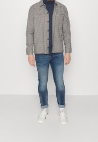 Ruudullinen nappipaita beige- ja mustavalkoisena tummansinisen poolopaidan päällä. Slim-fit siniset farkut ja valkoiset tennarit, joissa on punaisia yksityiskohtia.