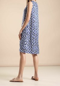 Bezramenné modro-bílé šaty s geometrickým vzorem a volně stojícím střihem, dosahující ke kolenům, v kombinaci s plochými sandály.
