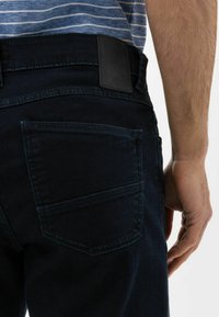 I jeans in denim scuro presentano una texture liscia, una vestibilità slim e uno stile distintivo delle tasche posteriori. Un'etichetta in pelle nera del marchio è attaccata al cinturino.