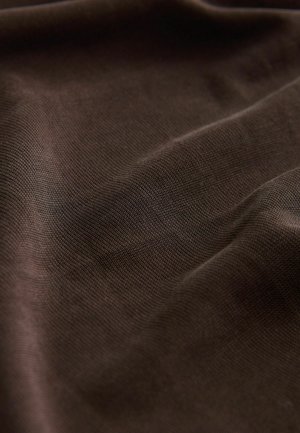 Tumma ruskea kangas hienolla, teksturoidulla kudonnalla, jossa on hienovaraisia laskoksia ja varjoja, mikä viittaa pehmeään ja muokkautuvaan materiaaliin.