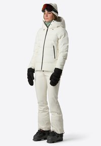 Biała kurtka puchowa z kapturem i zasuwanymi kieszeniami, w połączeniu z białymi spodniami narciarskimi i czarnymi rękawicami. Czarne buty narciarskie z metalowymi akcentami dopełniają cały look.